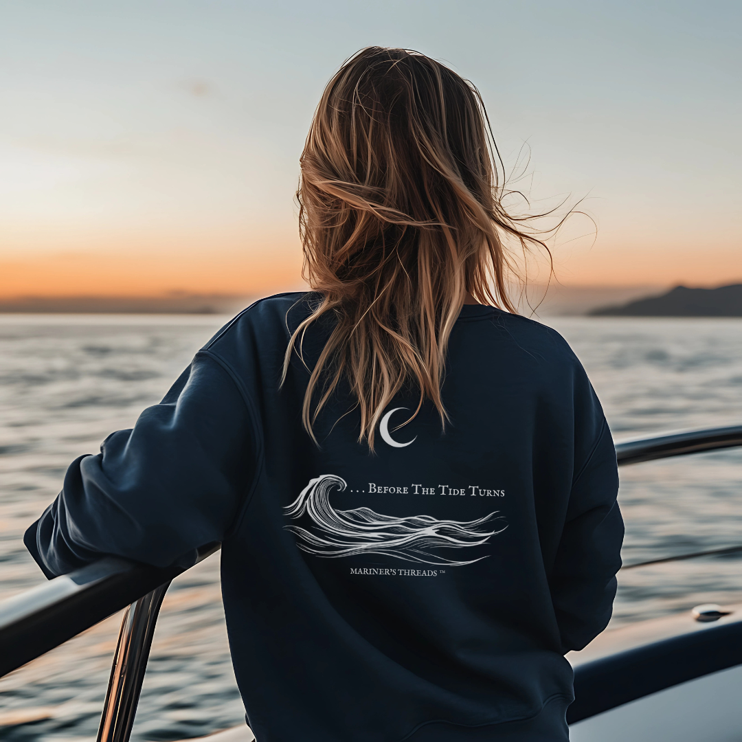 A woman with wavy blonde hair stands at the railing of a boat, gazing out at the open ocean during a breathtaking sunset. She is wearing a navy blue long-sleeve sweatshirt, showcasing a detailed back design featuring a crescent moon above stylized ocean waves, along with the phrase "...Before The Tide Turns" in an elegant, nautical font. The Mariner’s Threads™ brand name is subtly placed beneath the design.