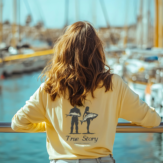 Woman with wavy brown hair leaning on a marina railing, looking out at sailboats while wearing a soft yellow Mariner’s Threads™ long-sleeve tee. The back of the shirt features a playful silhouette design of a fisherman holding a fish next to a woman holding a larger fish, with the phrase ‘True Story,’ capturing the humor and adventure of life on the water.