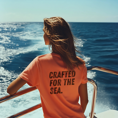 Woman standing at the railing of a boat, gazing at the open ocean with the wind in her hair, wearing a coral Mariner’s Threads™ T-shirt. The back of the shirt features the bold phrase ‘Crafted for the Sea,’ reflecting a passion for maritime adventure and coastal living.