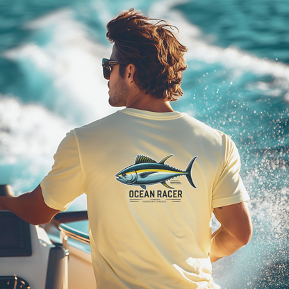  A sun-kissed man with tousled brown hair and sunglasses steers a high-speed boat, cutting through ocean waves. He wears a light yellow performance t-shirt, featuring a yellowfin tuna and the phrase "Ocean Racer" on the back, with Mariner’s Threads™ branding. The dynamic water spray and sunlight enhance the offshore racing and deep-sea fishing vibe.
