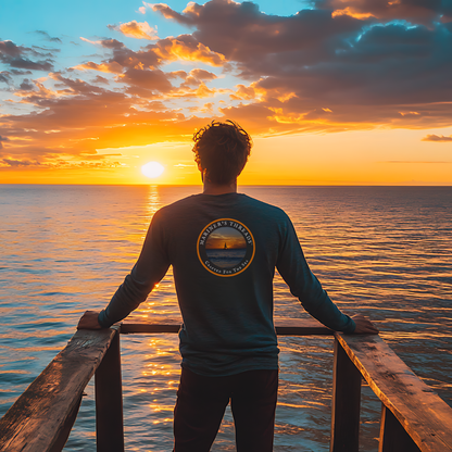 Man standing on a wooden pier at sunset, gazing at the horizon over the calm ocean while wearing a navy Mariner’s Threads™ long-sleeve tee. The back of the shirt features a circular sunset design with the brand's name and the phrase ‘Crafted for the Sea,’ capturing the essence of a life lived by the sea.