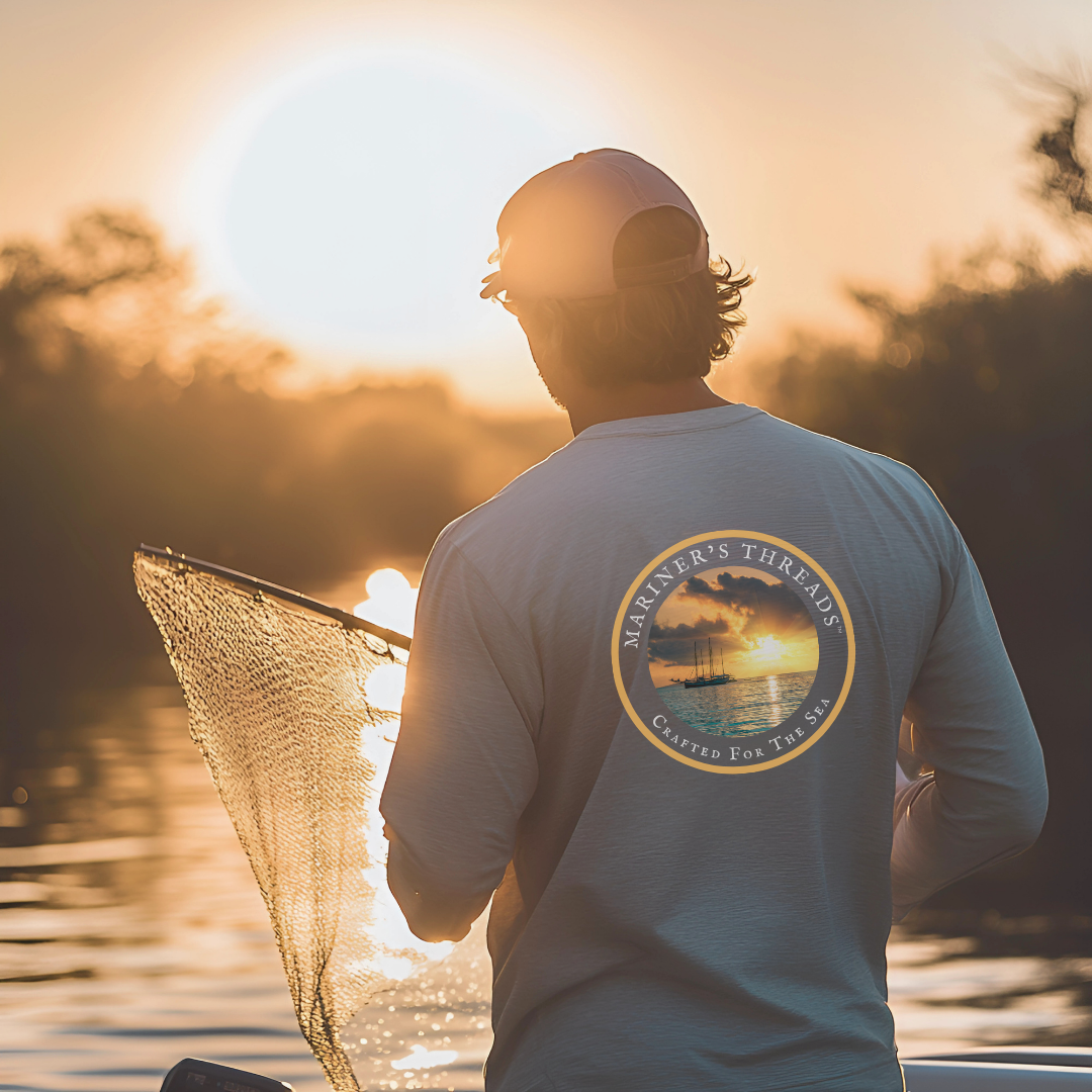 A rugged man wearing a light grey long-sleeve performance fishing shirt stands at the edge of a boat, casting a net into the calm waters at sunrise. The back of the shirt features a circular design with a stunning sunset over the ocean and the words "Mariner’s Threads – Crafted for the Sea" elegantly displayed around the border. The golden sunlight reflects off the water and the net, creating a warm, tranquil atmosphere. His cap shields his eyes as he focuses on the cast, embodying the essence of the early.
