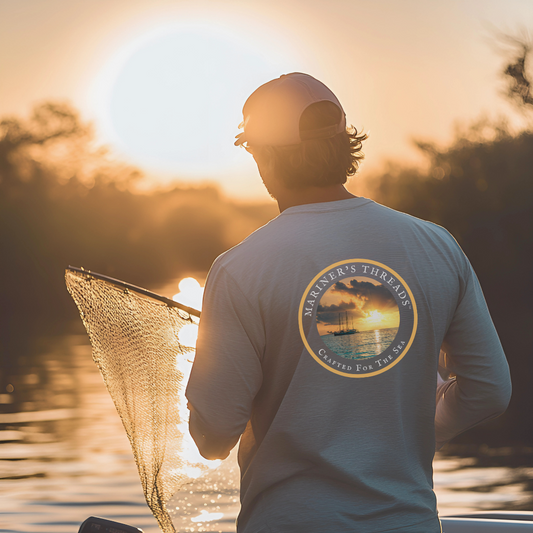 A rugged man wearing a light grey long-sleeve performance fishing shirt stands at the edge of a boat, casting a net into the calm waters at sunrise. The back of the shirt features a circular design with a stunning sunset over the ocean and the words "Mariner’s Threads – Crafted for the Sea" elegantly displayed around the border. The golden sunlight reflects off the water and the net, creating a warm, tranquil atmosphere. His cap shields his eyes as he focuses on the cast, embodying the essence of the early.